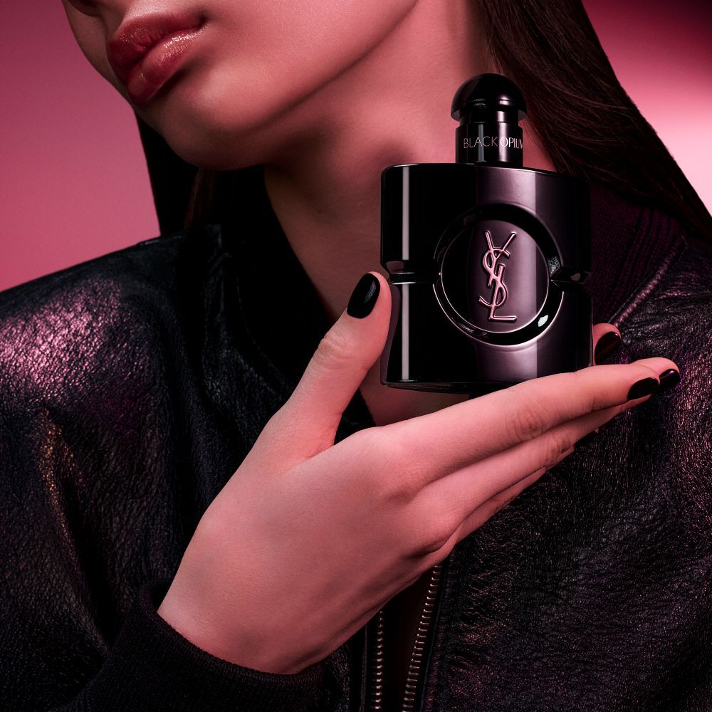 Black Opium Le Parfum bottle in glossy black, surrounded by dark vanilla pods and white jasmine with claims.