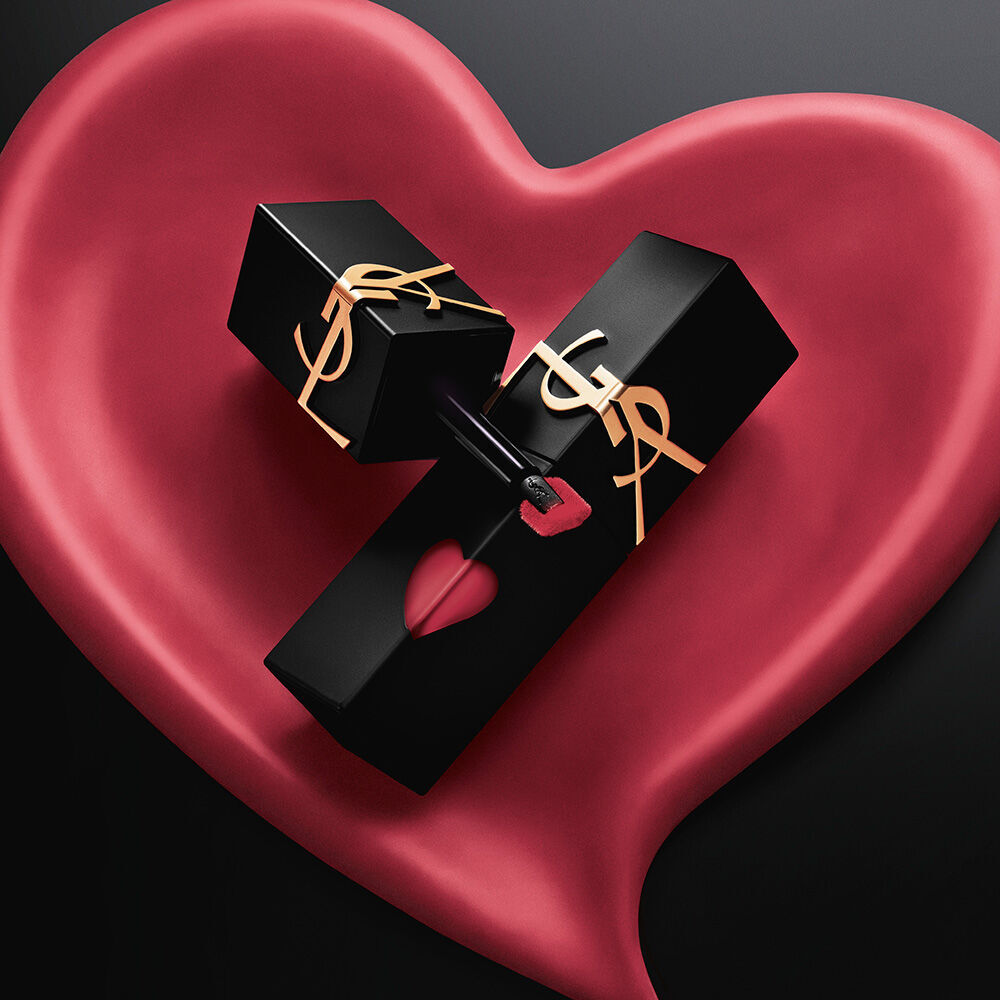 Two matte black YSL lip stain bottles with pink heart cutouts and gold logos, displayed inside a matching sculpted matte pink heart on a black background.