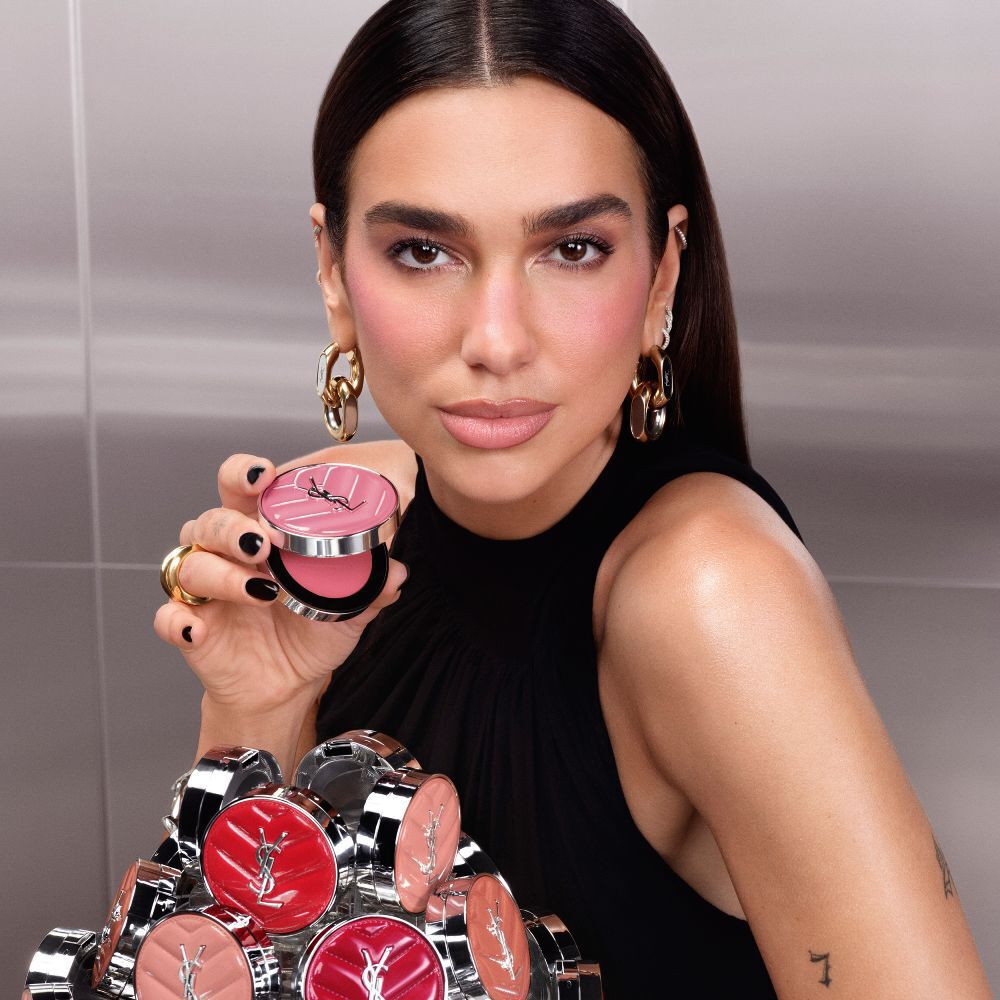 Dua Lipa holding an open YSL Make Me Blush Lifted Glow Cream Blush compact with a collection of the new shades displayed in front of her.