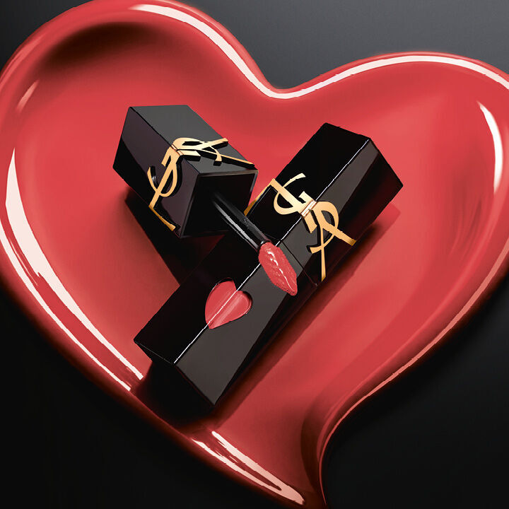 Two matte black YSL lip stain bottles with pink heart cutouts and gold logos, displayed inside a matching sculpted matte pink heart on a black background.