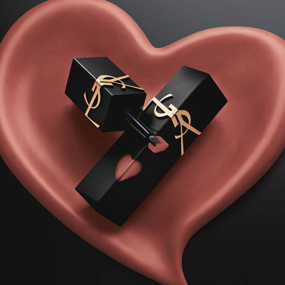 Two matte black YSL lip stain bottles with pink heart cutouts and gold logos, displayed inside a matching sculpted matte pink heart on a black background.