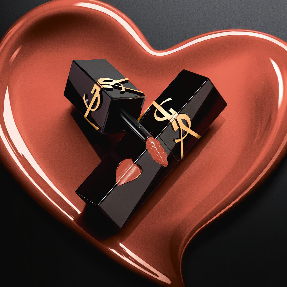 Two matte black YSL lip stain bottles with pink heart cutouts and gold logos, displayed inside a matching sculpted matte pink heart on a black background.