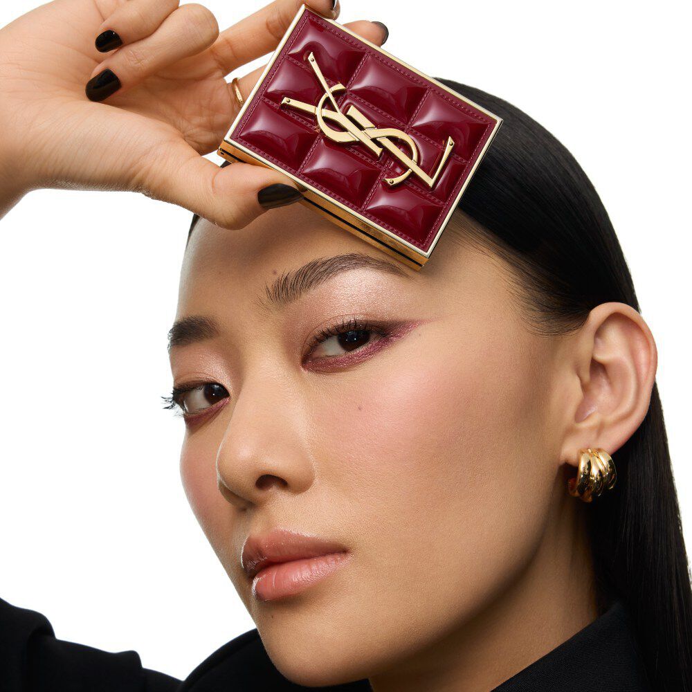 Model wearing a black suit and glam look, seated in a festive candlelit setting with YSL gift boxes.
