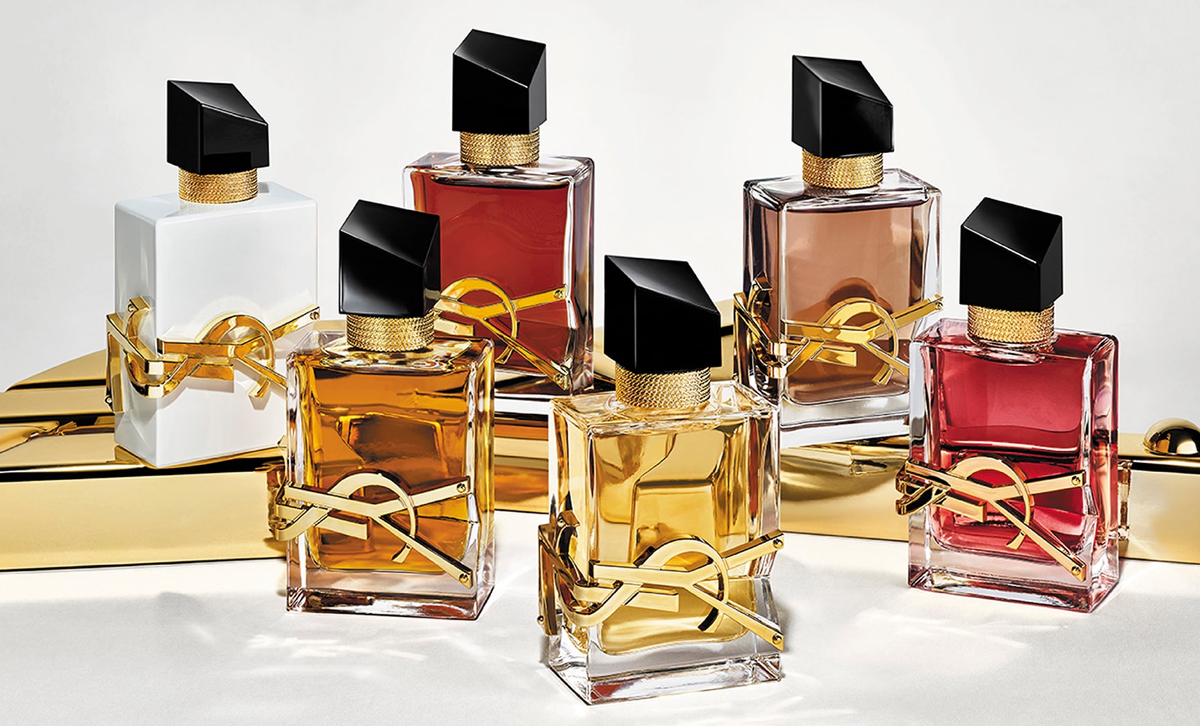 Six YSL Libre perfume bottles displayed on a shiny gold surface. The bottles show variations in the color of the liquid and the bottle finish, including clear, white, reddish and brown hues. All feature the iconic gold YSL lettering and a slanted black cap.