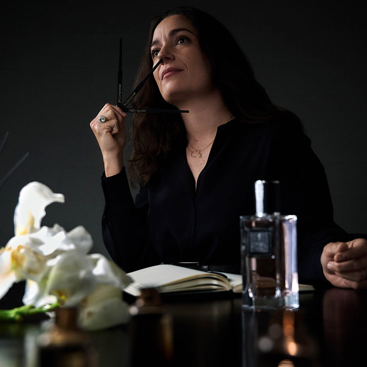 A woman in a black shirt holds a perfume blotter, looking up thoughtfully, with a notebook, perfume bottle, and flowers on the table.