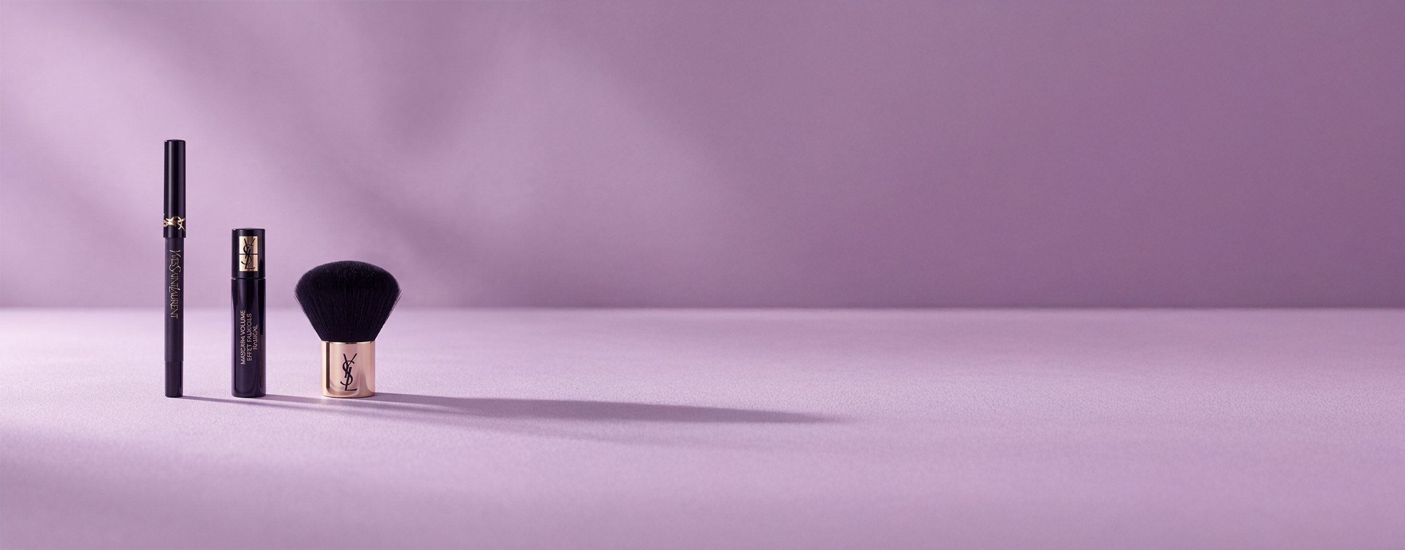 Three Yves Saint Laurent beauty products, including a mascara and a kabuki brush, on a soft lavender background.