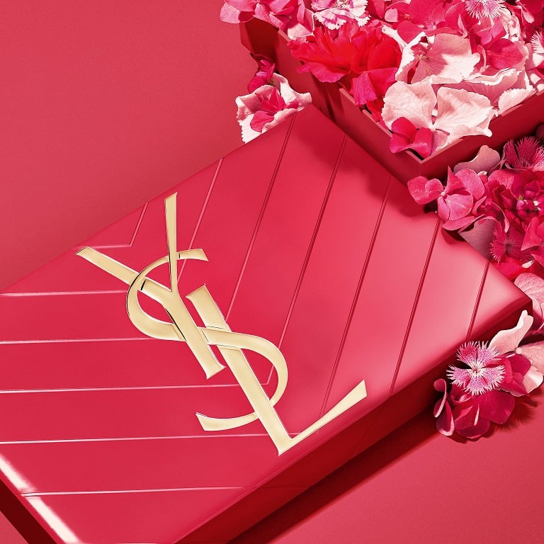 Coffret cadeau YSL rose vif avec logo Cassandre doré, entouré de pétales de fleurs printanières.