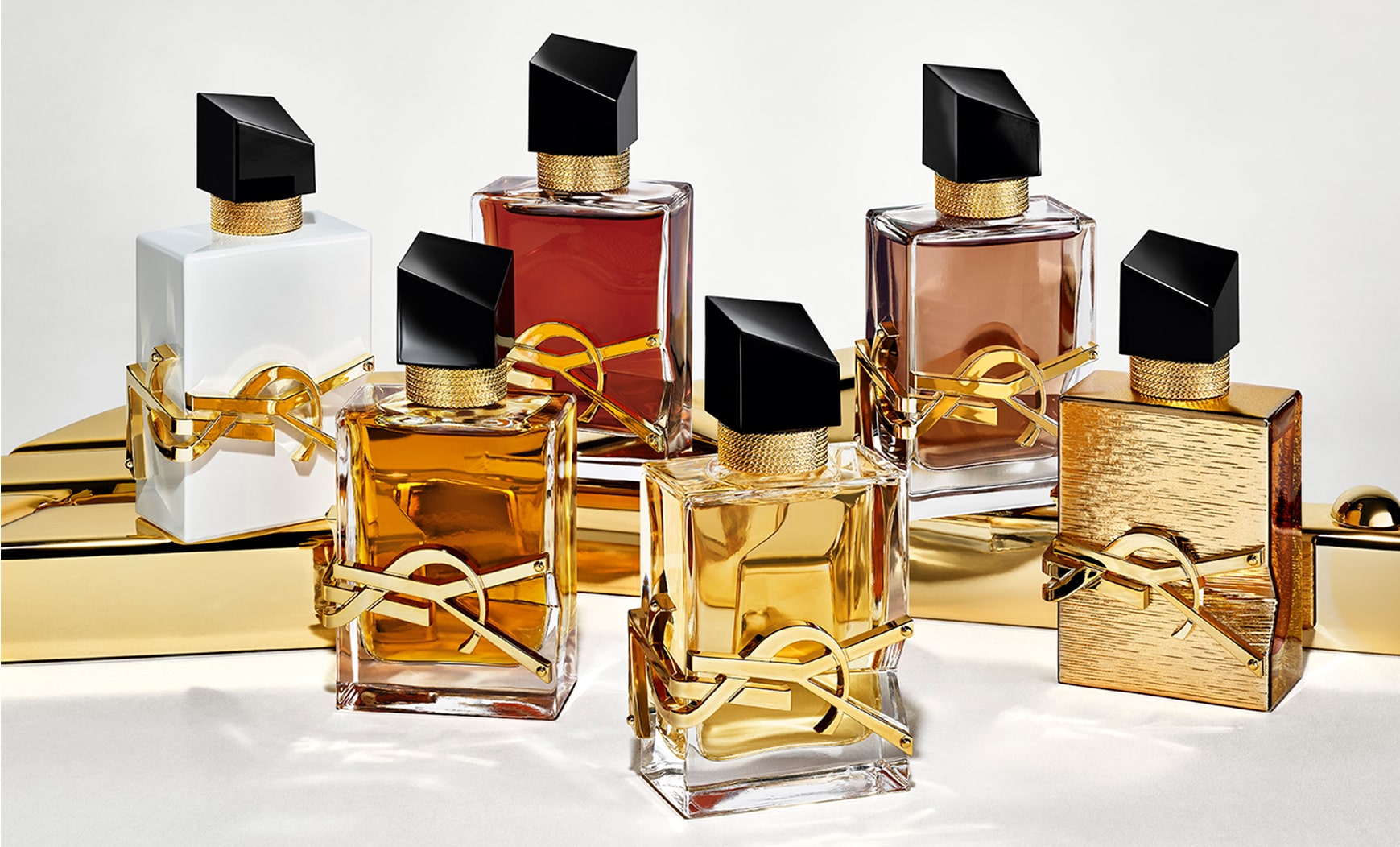 Six YSL Libre perfume bottles displayed on a shiny gold surface. The bottles show variations in the color of the liquid and the bottle finish, including clear, white, reddish and brown hues, and a textured all-gold bottle. All feature the iconic gold YSL lettering and a slanted black cap.