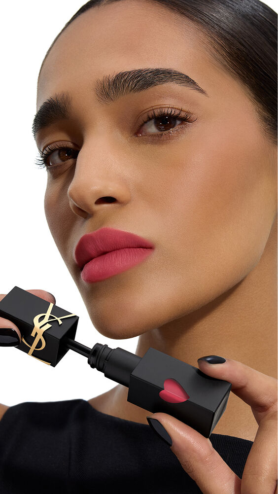 Medium skin tone model with a vibrant rose-matte lip color holding the black YSL THE INKS BLUR lip stain bottle with its pink heart.