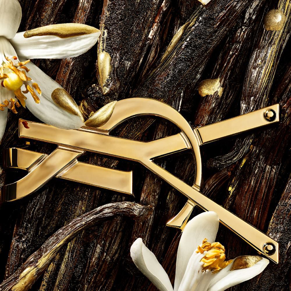 A golden YSL logo sits atop a bed of dark, textured vanilla pods and white orange blossoms, accented with gold flakes.