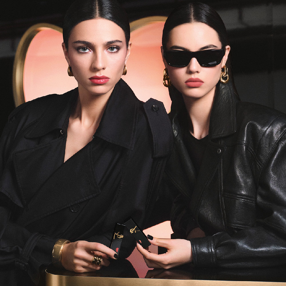 Two chic models in black trench and leather jackets holding YSL lipsticks in front of a large pink heart-shaped backdrop, showcasing a glossy red lip look and a soft-matte pink lip look.