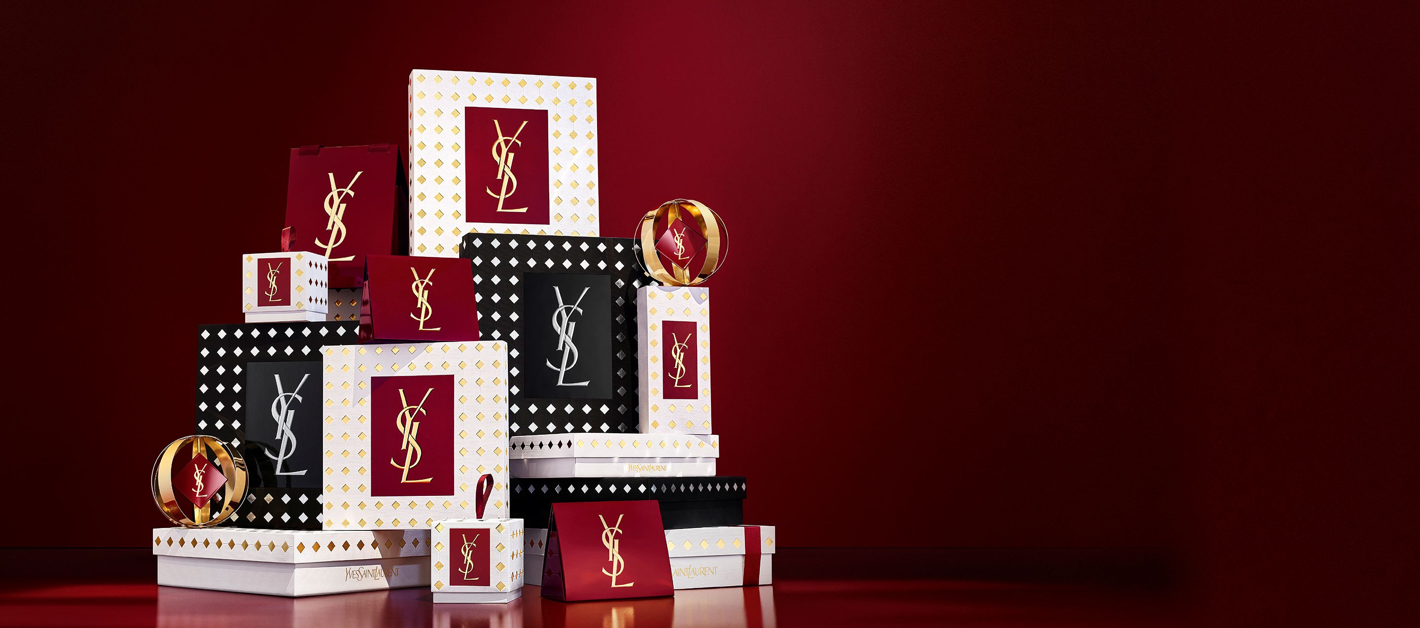 Three models in elegant black outfits holding YSL holiday gift boxes in front of a festive red curtain background with falling gold confetti.