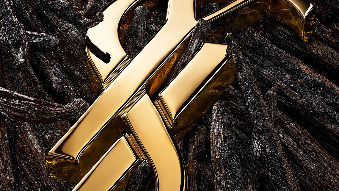 A golden YSL logo sits atop a bed of dark, textured vanilla pods and white orange blossoms, accented with gold flakes.