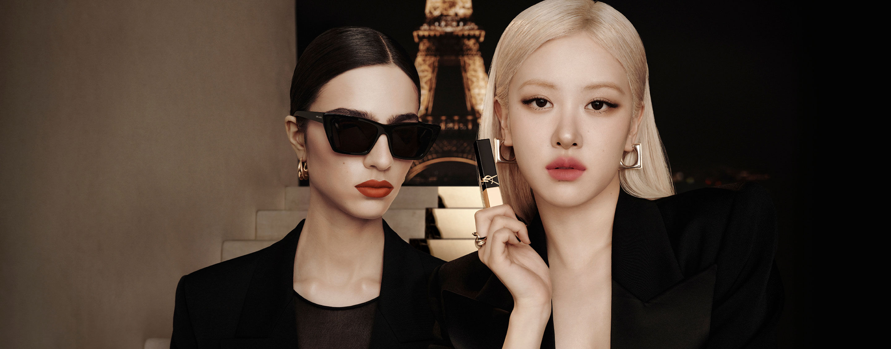 Two models in black before the Eiffel Tower at night; one holds a gold-and-black YSL lip product.