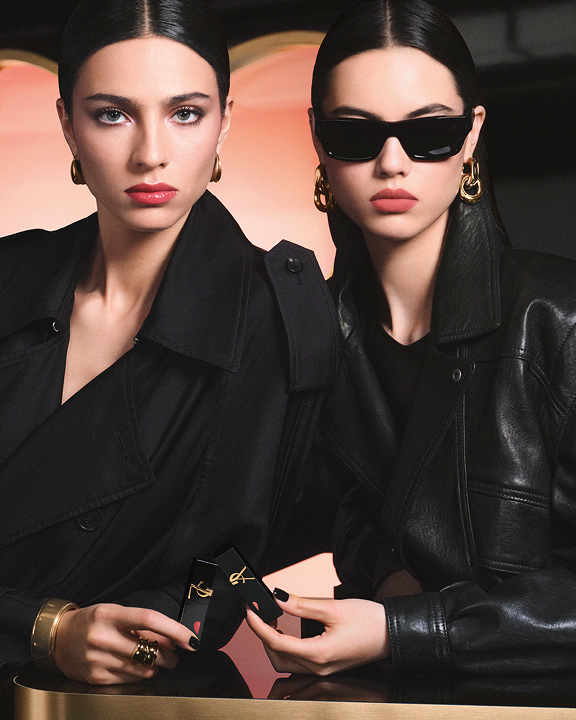 Two chic models in black trench and leather jackets holding YSL lipsticks in front of a large pink heart-shaped backdrop, showcasing a glossy red lip look and a soft-matte pink lip look.