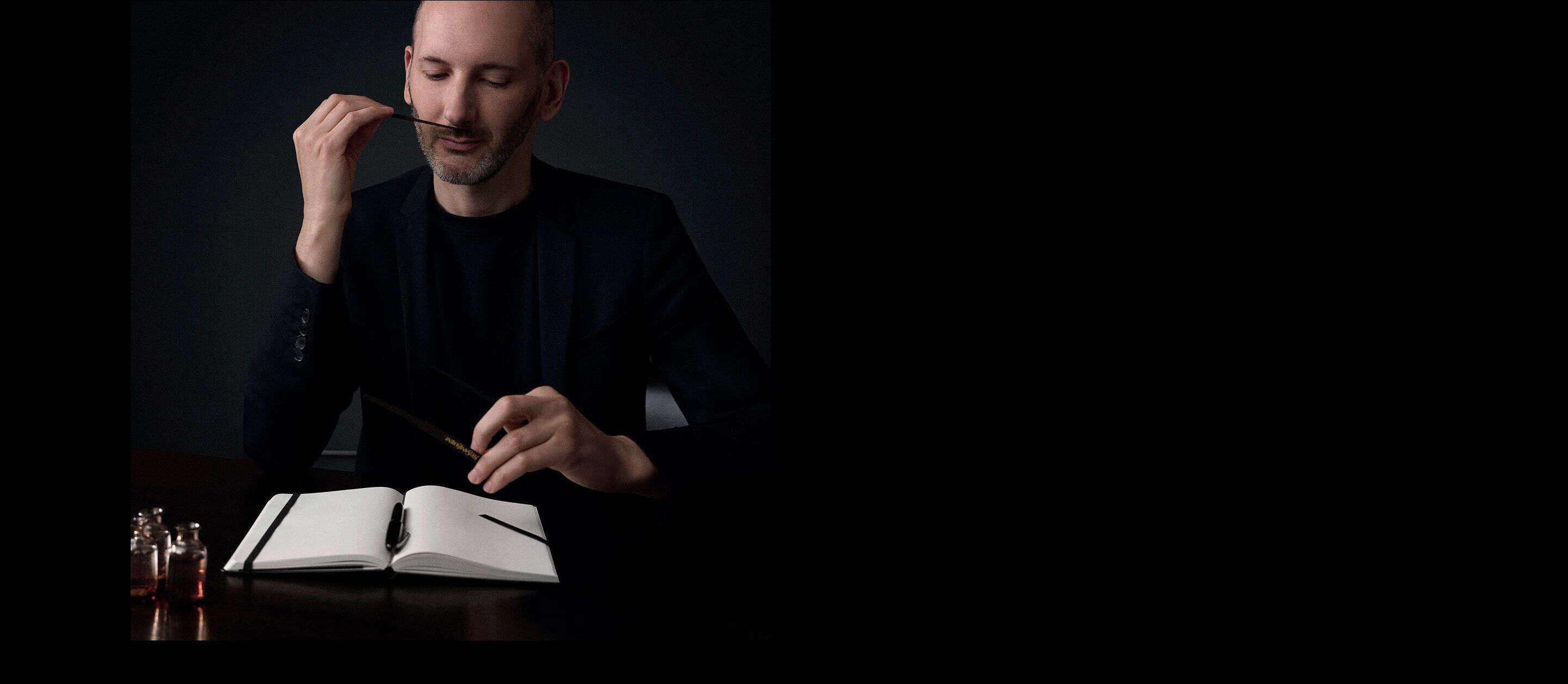 A perfumer in a dark suit smelling a fragrance strip while working at a desk with an open notebook and small perfume vials.