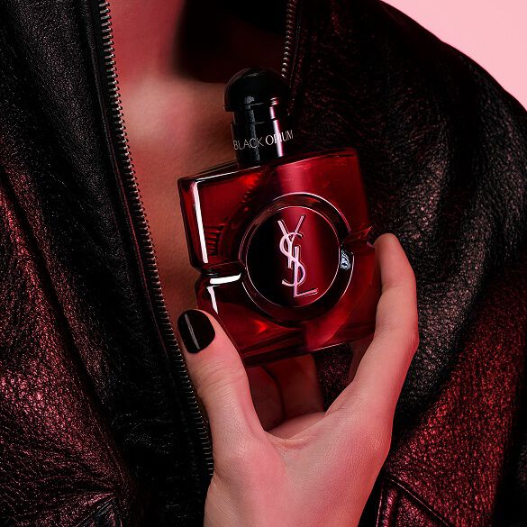 Close-up of a hand with black nails holding the Black Opium Eau de Parfum Over Red bottle against a black leather jacket.